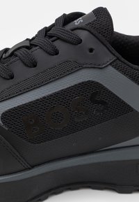Sneaker noire avec un mesh texturé et des matériaux lisses, dotée de lacets noirs et d'un logo "BOSS" en bold sur le panneau latéral.