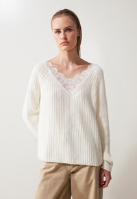 Witte gebreide trui met een geribbelde structuur, V-hals ontwerp met een kanten rand. Het model draagt beige broek. De achtergrond is licht en neutraal.
