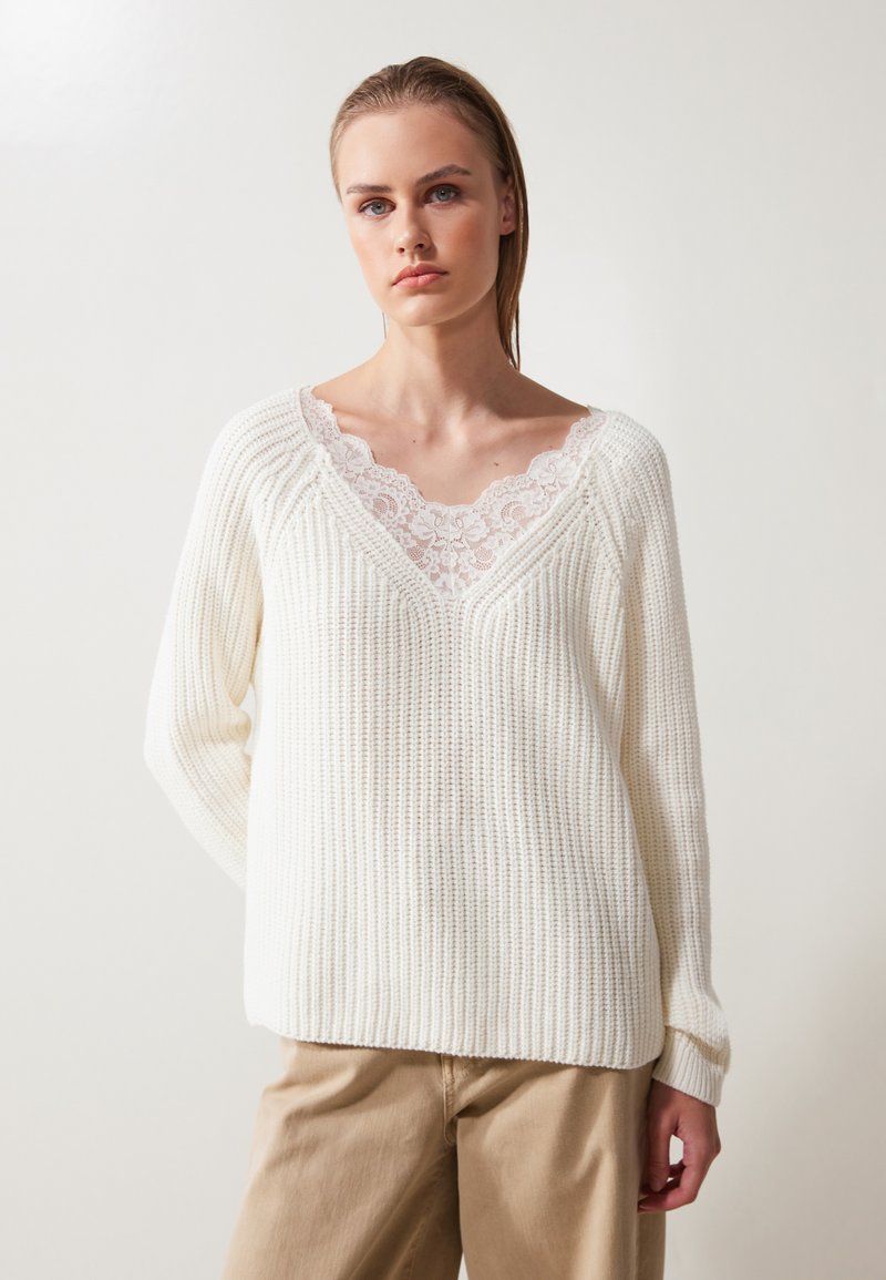 Pull en maille blanche avec une texture côtelée, design en V avec un volant en dentelle. Le modèle porte un pantalon beige. L'arrière-plan est clair et neutre.