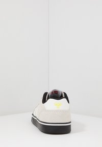 Veloursgraue Sneaker mit schwarzen Akzenten, weißer Ferse, gelbem Logo-Detail an der Ferse und Gummisohle. Minimalistisches Design mit niedrigem Profil.