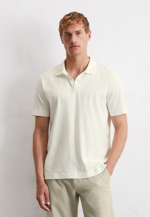 Polo à manches courtes en coton écru avec col à revers, coupe droite et fentes latérales ; associé à un pantalon beige clair.