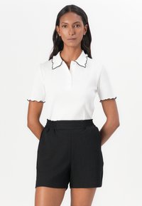 Polo shirt bianco con colletto nero scallop e maniche corte, abbinato a pantaloni corti neri a vita alta con texture.