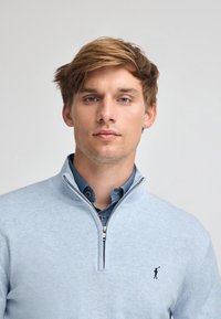 Ljusblå zip-tröja med slät textur och ribbad krage, lagd över en mörkblå skjorta med krage. Har loggan broderad.