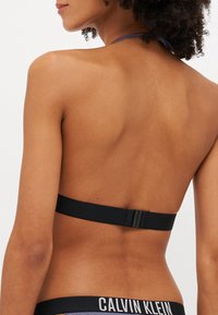Černá podprsenka s halter střihem a zapínáním na zadní straně. Hladká tkanina a ramínka zdůrazňují jednoduchý design. Viditelné logo Calvin Klein.