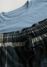 Hellblau gestreiftes Langarmshirt, kombiniert mit schwarzer und beiger Karohose mit Kordelzug, die aus einem strukturierten Stoff gefertigt ist und einen elastischen Bund hat.