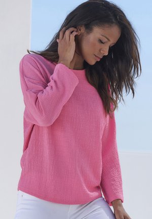 Pull en maille rose clair avec manches longues, coupe ample et col rond, associé à un pantalon blanc.