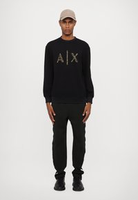Sudadera negra con el logo dorado "A|X", combinada con pantalones verde oscuro y zapatos negros. El modelo lleva una gorra de béisbol beige.