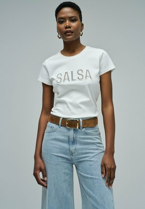 Mujer con una camiseta blanca con el texto "SALSA", pantalones vaqueros de talle alto en azul claro y un cinturón de ante marrón con hebilla dorada.