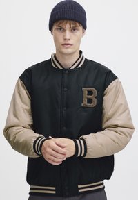 Black and beige bomber jacket with a textured letter "B" patch, snap buttons, and striped ribbed cuffs and hem. Worn over a gray shirt.