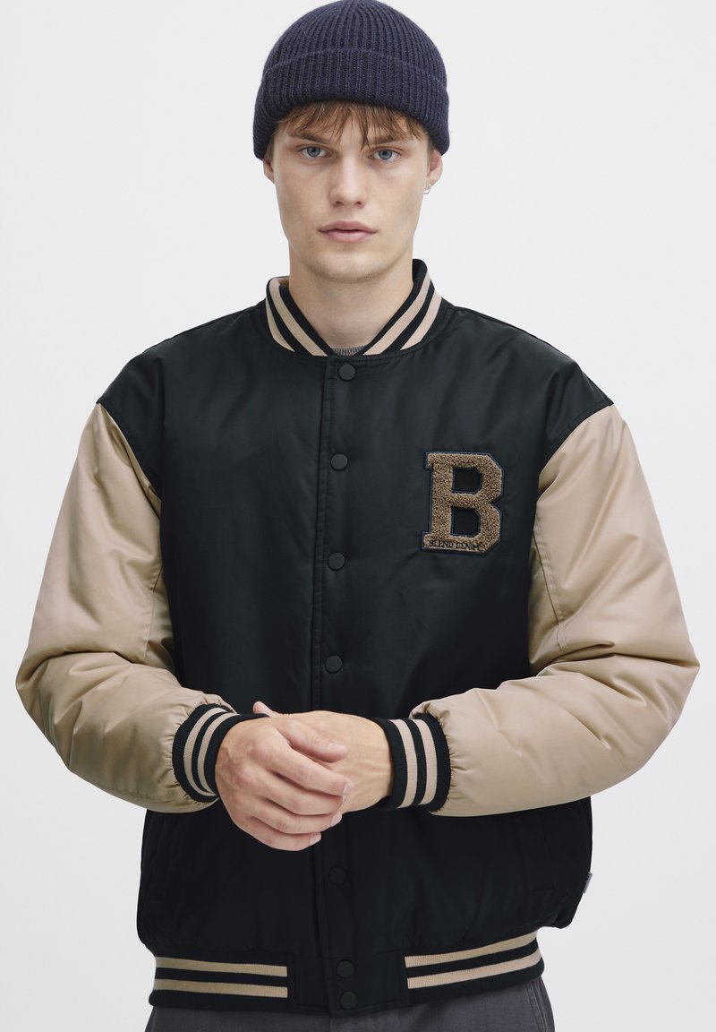 Black and beige bomber jacket with a textured letter "B" patch, snap buttons, and striped ribbed cuffs and hem. Worn over a gray shirt.