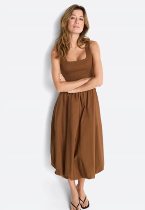 Žena stojaca so založenými rukami, oblečená v hnedých midi šatách bez rukávov a čiernych žabkách, na bielom pozadí.