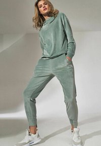 Groene velours trainingsoutfit met een losse hoodie met hoge kraag en taps toelopende broeken met zijzakken, gecombineerd met witte sneakers.