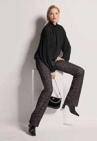 Schwarze Bluse mit langen Ärmeln und gerafftem Ausschnitt, kombiniert mit Hosen im Tweed-Muster und weitem Bein. Schwarze Handtasche aus Lackleder mit Kettenriemen. Stiefeletten mit Absätzen.