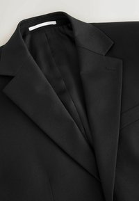 Veste de costume noire sur mesure avec revers crantés, couture apparente sur la boutonnière et texture de tissu lisse sur fond clair.