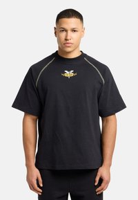 Sort bomulds T-shirt med korte ærmer, ribbet halsudskæring og guld logo detaljer på brystet. Har kontrasterende syninger langs skuldrene.