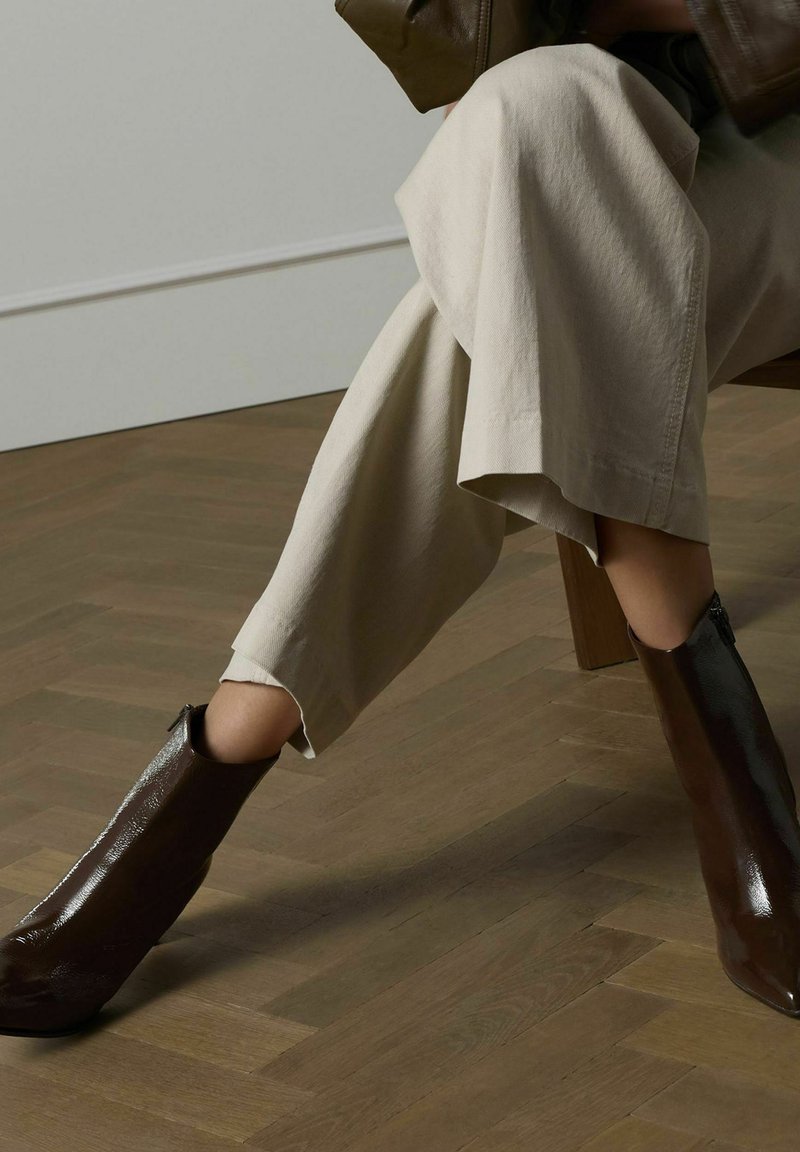 Brown patent leather ankle boots with a tapered toe and a side zipper, paired with wide beige trousers, set against a wooden floor.