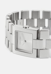 Montre bracelet rectangulaire en argent avec un cadran en métal brossé, au design minimaliste, et un bracelet en métal composé de maillons carrés.