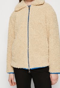 Bežová fleecová bunda s texturovaným, plyšovým povrchem, s límcem, předním zipem a modrým podšívkou na rukávech a dolním lemu.