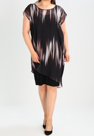 Vrouw die een zwarte jurk draagt met een doorschijnende, asymmetrische overlay met verticale witte en bruine strepen, gecombineerd met zwarte hakken.