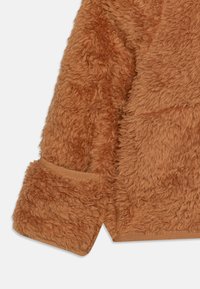 Tessuto in pile marrone e soffice, con una superficie strutturata. Include una tasca semplice e integrata e un dettaglio del polsino liscio.