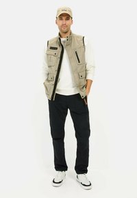 Beige Utility-Weste mit mehreren Taschen, schwarzen Reißverschlüssen, kombiniert mit einem weißen Langarmshirt und schwarzen Hosen, abgerundet mit weißen Sneakern.