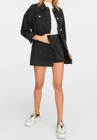 Frau in schwarzer Jeansjacke und -rock, weißem Hemd, die ihren Rock anpasst, steht mit weißen Sneakers vor einem neutralen Hintergrund.