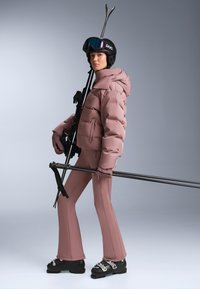 Skijaško odijelo koje uključuje ružičastu jaknu na naduvavanje i hlače u istoj boji, s crnim skijaškim čizmama i štapovima. Značajni su teksturirana tkanina i prianjajući kroj.