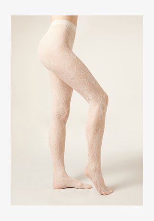 Femme portant des collants en dentelle nude transparente avec un motif floral, debout sur la pointe des pieds sur un fond clair et uni.