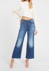 Helle Blue-Jeans mit weitem Bein, leichter Ausbleichung, fransigem Saum und hoher Taille. Kombiniert mit einem kurz geschnittenen, gerippten weißen Pullover.