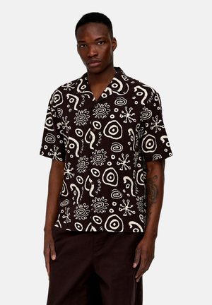 Homme portant une chemise noire à manches courtes avec des motifs abstraits blancs circulaires et ondulés, se tenant devant un fond uni.