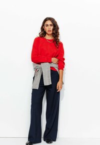 Sweatshirt rouge au fit décontracté, pull gris noué à la taille, pantalon large à rayures bleu marine, et chaussures noires à pois blancs.
