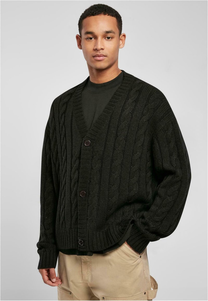 Svart kabelstrikket cardigan med dyp V-hals, lange ermer og brune knapper, lagt over en mørk skjorte, med en avslappet passform.