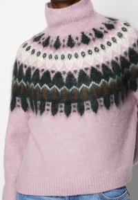 Różowy, wełniany sweter z golfem z geometrycznym wzorem w ciemnozielonym, białym i brązowym kolorze. Miękka faktura, krótki fason z prążkowanym wykończeniem na dole.