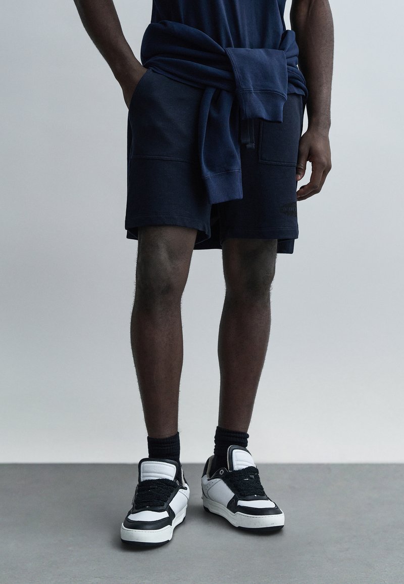 Σκούρα μπλε σορτς με υφή, σε συνδυασμό με μαύρα και λευκά αθλητικά παπούτσια. Μπλε σκούρο φούτερ είναι δεμένο γύρω από τη μέση.