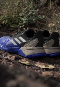 Mustat ja siniset urheilukengät vedenpitävällä materiaalilla, jossa on teksturoitu pinta, valkoiset raidat ja vaaleanruskea yksityiskohta kannassa.