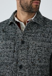 Kabát s vzorom rybej kosti v čiernej, bielej a hnedej farbe. Obsahuje zapínanie na gombíky, golier a textúrovanú tkaninu s viditeľnými švovými detailmi.