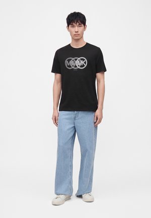 Jeune homme portant un t-shirt graphique noir, un jean droit bleu clair et des baskets blanches, debout sur un fond blanc uni.