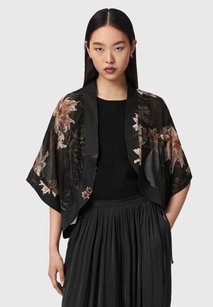 Vrouw draagt een zwarte geplooide rok, een zwarte top en een doorschijnend zwarte bloemrijke kimono-stijl jas met brede mouwen.