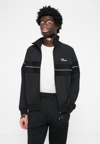 Sort zip-up jakke med hvide striber og "Ellesse" logo. Kombineret med sorte bukser med elastisk talje. Glat stofstruktur.