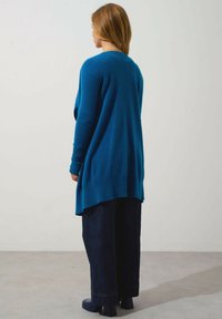 Cardigan blu in maglia con maniche lunghe, polsini e orlo a coste, indossato sopra jeans larghi scuri. Tessuto liscio e vestibilità comoda.
