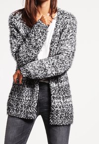 Texturerad svartvit cardigan med lös passform, öppen front och långa ärmar, tillverkad av mjukt och lurvigt material. Bärs över en vit topp.