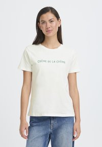 Kremno-bel bombažni majica s kratkimi rokavi. Na prsih je zeleni napis "CRÉME DE LA CRÉME". Ohlapen kroj, minimalističen dizajn.