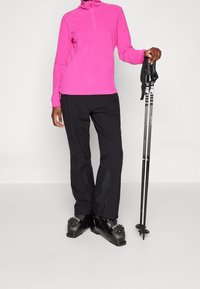 Růžový fleecový svetr na zip, černé lyžařské kalhoty s texturovanými panely a černé lyžařské boty. Drží lyžařské hůlky s rukojeťmi s logy značky.