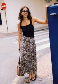 Frau trägt schwarzen Tanktop, Leopardenmuster-Rock, Sonnenbrille und Flip-Flops, lächelt und hält einen blauen Briefkasten auf einem Bürgersteig in der Stadt.