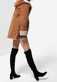 Abrigo de borrego marrón con capucha, forro crema y corte ligeramente oversize, combinado con botas negras por encima de la rodilla con detalle arrugado.