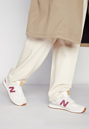 Osoba w beżowych spodniach i białych butach New Balance z bordowym logo stoi na jasnoszarej podłodze na tle jednolitej ściany.