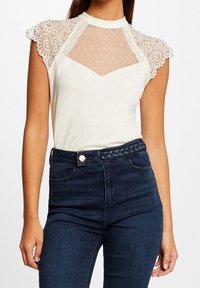 Hvid top med blonde-kappelærmer og et gennemsigtigt, prikket panel, kombineret med mørkeblå jeans med flettet bælte.