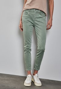 Vaqueros ajustados de mezclilla de color verde menta, confeccionados en una mezcla de algodón, que cuentan con cinco bolsillos, detalles de costura y largo hasta el tobillo, combinados con zapatillas blancas.