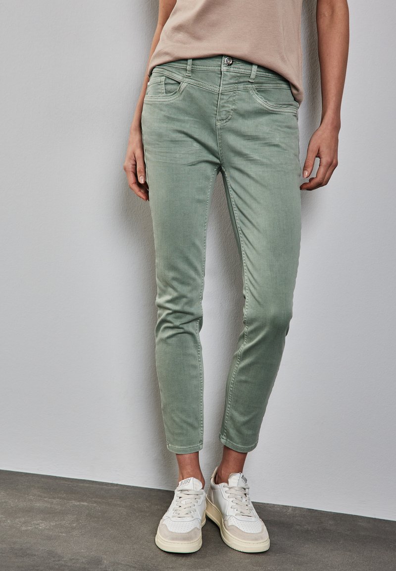 Vaqueros ajustados de mezclilla de color verde menta, confeccionados en una mezcla de algodón, que cuentan con cinco bolsillos, detalles de costura y largo hasta el tobillo, combinados con zapatillas blancas.