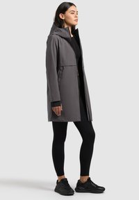 Ženska, ki nosi sivo kapucarasto nepremočljivo jakno srednje dolžine čez črne legice in črne superge, stoji v profilu na enostavni ozadju.